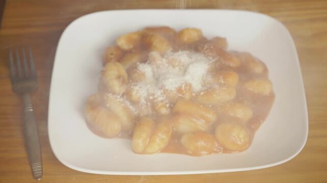 Grated parmesan cheese falling over a plate of hot steaming gnocchi pasta, Close up of sprinkling cheese onto delicious potato gnocchi with red sauce