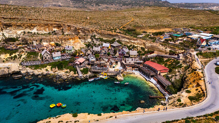 aerial drone view of Popeye village, malta on anchor bay coast on Mediterranean Sea in summer with clear blue sunny skies overhead and boats below with Woden houses from old movie set theme park © STJMEDIA
