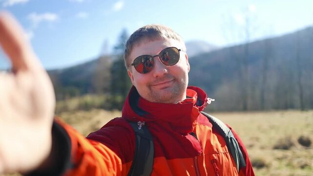 A close-up shot of a cheerful Caucasian male traveler with a backpack and orange jacket smiling and taking a selfie during a mountain hike on a sunny day.