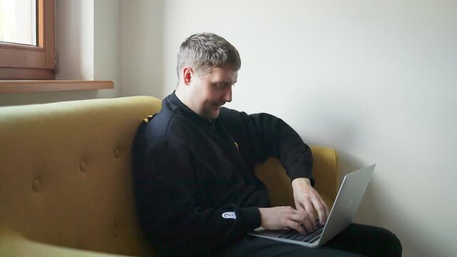 A side view of a young Caucasian man sitting on a modern yellow couch, holding his head thoughtfully while working on a laptop computer in a bright minimalist room.