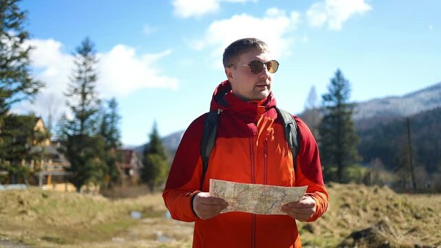 A young handsome male traveler in an orange jacket and sunglasses holding a paper map and pointing away on a sunny day during a mountain hike.
