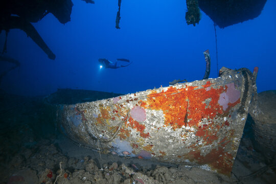 Egypt - 07 May 2021: View of a sunken shipwreck covered in colorful coral growth on the seabed with a scuba diver exploring the site with a flashlight in the deep blue water.