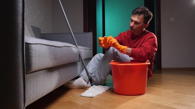 Man wearing orange protective gloves and red sweatshirt, feeling exhausted while sitting on the floor after mopping with a bucket of water and a mop, taking a break from housework