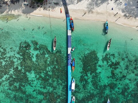 Aerial view of the floating pier at Ko Racha Yai with longtail boats moored in turquoise waters, sandy beach, and coral reefs in Rawai, Phuket, Thailand.