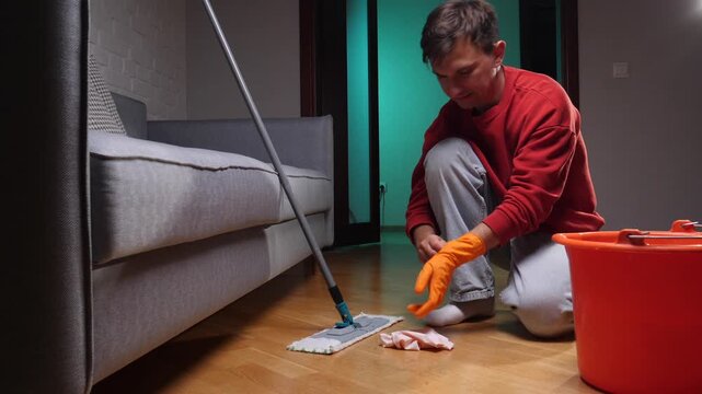 Man in casual clothing cleaning the wooden floor of his living room with a mop and an orange bucket, performing daily chores and maintaining a tidy home