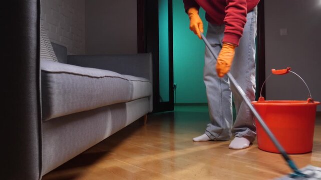 Person wearing orange protective gloves is actively mopping the wooden floor in a living room, performing regular household chores and maintaining cleanliness at home