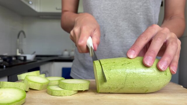 Woman slicing fresh green zucchini on a wooden cutting board in kitchen. Healthy food preparation.