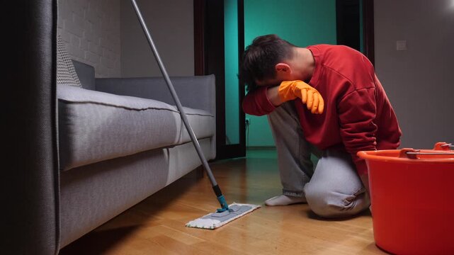 Man wearing an orange hoodie and gloves is kneeling on the floor, resting his head on his hand while holding a mop, feeling tired after cleaning the house