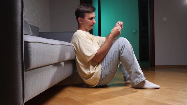 Young man sitting on the floor leaning against a sofa, experiencing frustration and annoyance while trying to unravel a messy knot of wired earphones indoors