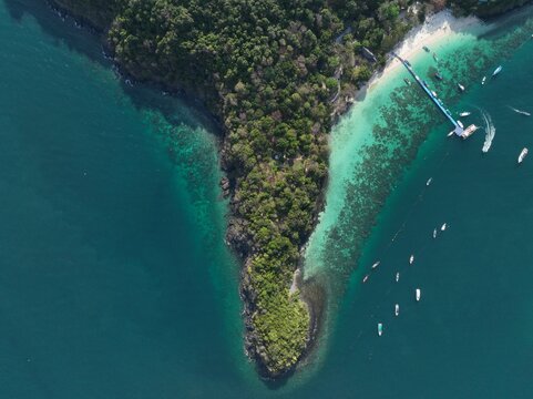 Aerial view of Ko Racha Yai with lush tropical vegetation, turquoise coral reef waters, a wooden pier, and small boats anchored in the Andaman Sea, Rawai, Phuket, Thailand.