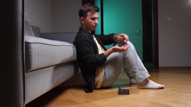 Man sitting on the living room floor next to a couch, holding an empty wallet and extending his hand, symbolizing financial struggle, poverty, and debt