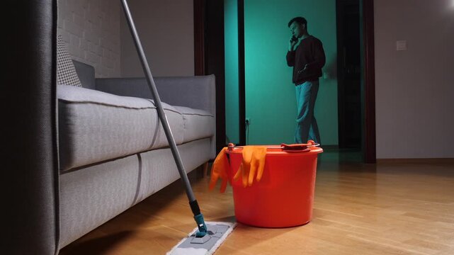Cleaning supplies including a mop, bucket, and orange gloves are on a wooden floor in a living room, with a man talking on a mobile phone in the background, ignoring chores