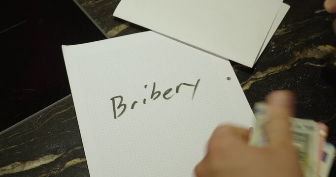 Close-up of a person writing the word "bribery" on paper and placing cash beside it. Concept of corruption, illegal payments, financial crime, and unethical business practices.