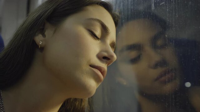 Young Caucasian woman resting her head against a rain-streaked window on a train, with soft lighting illuminating her serene expression and reflection visible in the glass