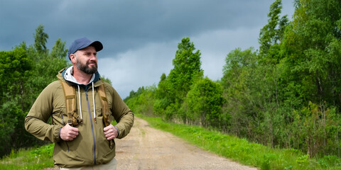 Bearded man with backpack stands on rural road surrounded by green forest and looks into distance before continuing route. Concept of independent travel © Dmitrii Masquerade