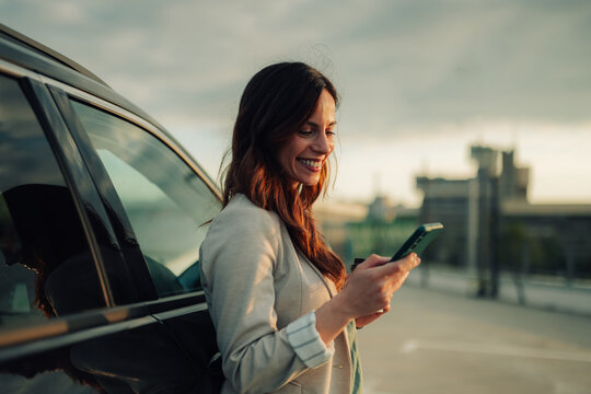 Woman standing by her car, smiling while texting on a smartphone, enjoying sunset light. Connecting on the go
