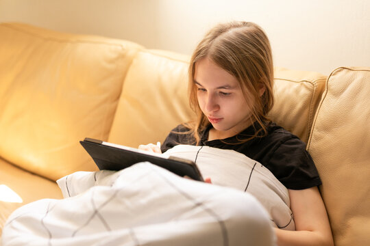 Blonde teenage girl using digital tablet and stylus while covered with blanket on sofa.