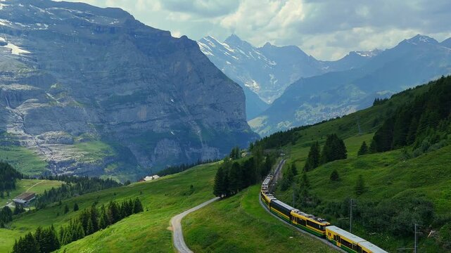 A wide, scenic landscape video showing a yellow and green passenger train traveling down a rack railway track cut into a lush green hillside, with a winding dirt road and a small pond in the foregroun