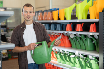 In garden hypermarket, guy carefully examines product before buying, chooses watering can for gardening. Amateur gardener selects necessary items tools for hobby in greenhouse flower-bed © JackF