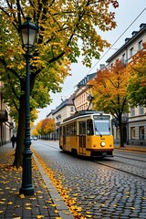 Obraz premium Yellow tram moves on cobblestone street with autumn leaves. Trees with yellow and orange foliage line the road. Buildings show classic European architecture, a single person walks on sidewalk.
