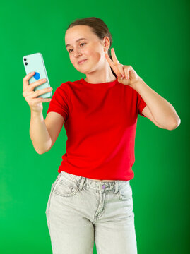 Teenage girl in red T-shirt and jeans takes selfie on a smartphone on a green background. Chroma key compositing