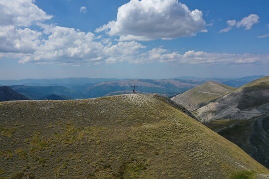 Aerial view of the summit of Pizzo Berro featuring a cross on the grassy peak with distant mountain ranges under a blue sky in Montefortino, Marche, Italy.
