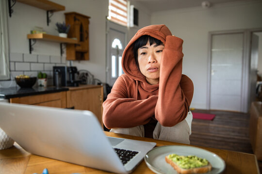 Stressed woman in hoodie working on laptop at home kitchen