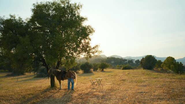 A man dismounts his horse and ties it to a tree trunk at sunset in countryside