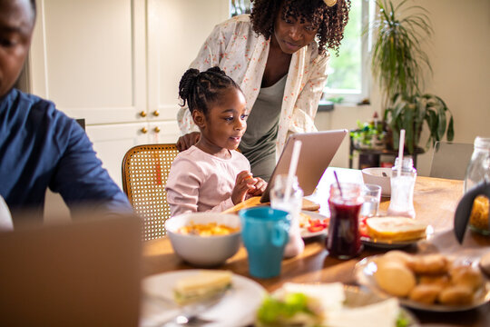 Mother helping daughter use tablet at breakfast in home kitchen