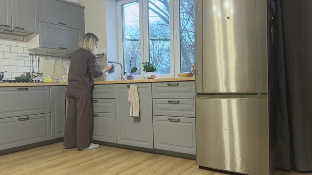 Woman cleaning kitchen faucet with cloth near sink. Everyday kitchen hygiene and home cleaning routine.