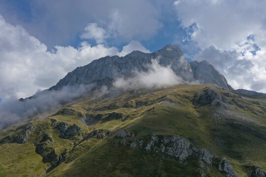 Aerial view of Corno Piccolo in the Gran Sasso massif, featuring rugged limestone peaks, grassy slopes, and wispy clouds clinging to the mountainside in Pietracamela, Abruzzo, Italy.