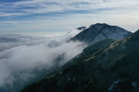 Aerial view of Monte Baldo with rugged mountain slopes partially covered by a thick layer of clouds under a clear blue sky in Malcesine, Veneto, Italy.