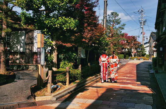 Kanazawa, Japan - 07 November 2025: View of two women in traditional kimonos walking along a historic stone-paved street in Kanazawa, surrounded by autumn foliage and traditional wooden architecture.