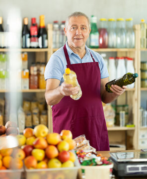 Elderly male seller at counter offering olive or sunflower oil in bottle in grocery store