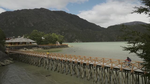 Person in a yellow jacket walks along a wooden boardwalk in the remote coastal village of Caleta Tortel, Chile, surrounded by scenic mountains and a bay.