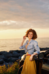 Fototapeta premium Young woman with curly hair stands beside a vintage car at sunset, wearing a denim shirt and bright yellow skirt, smiling warmly for the camera.