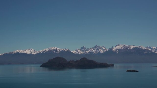 Wide landscape view of General Carrera Lake in Patagonia, featuring an island in the foreground and a prominent range of snow-capped Andes mountains under a clear sky.