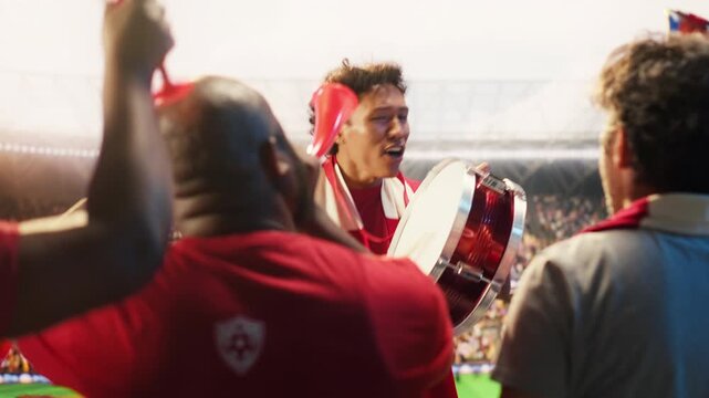Enthusiastic man in stadium beating drum, encouraging team and creating loud match day energy, showing players, showing football fan leader galvanizing red shirt supporters in live sports arena.