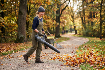 Naklejka premium Female worker using leaf blower in autumn park path