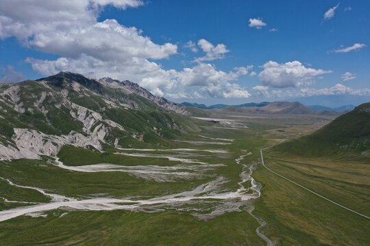 Aerial view of Campo Imperatore, a vast high-altitude plateau with rugged mountain slopes, green meadows, and a winding road under a blue sky in L'Aquila, Abruzzo, Italy.