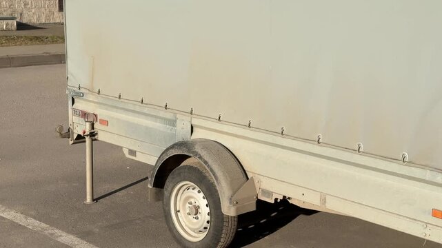 Covered passenger trailer parked in lot during spring sunlight gray tarpaulin fastened over cargo bay single axle wheel, hitch