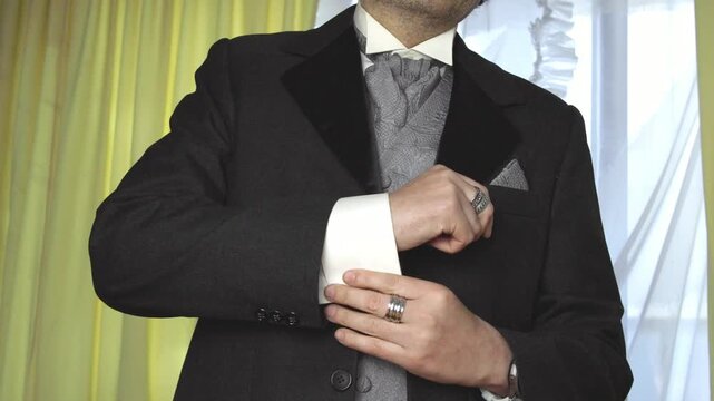Groom putting on elegant black suit jacket and adjusting gold cufflinks close up