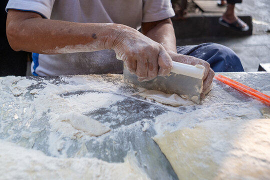 A street food vendor preparing cakwe (you tiao) dough with a metal bench scraper on a floured worktable at a traditional market in Central Java, Indonesia.