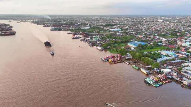 Aerial view of the Martapura River at Dermaga Banjar Raya with coal barges and small boats near the urban waterfront in Kecamatan Banjarmasin Barat, Kalimantan Selatan, Indonesia.