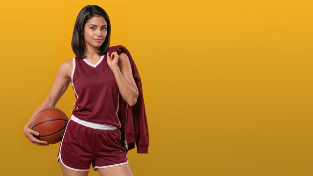 Young woman in burgundy basketball apparel holds a ball and drapes a matching jacket over her shoulder against a mustard yellow studio background with broad copy space.