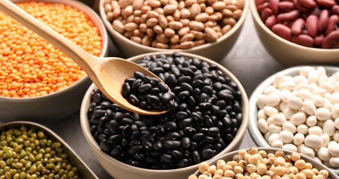 Taking black beans with spoon from bowl, different types of legumes on grey table, closeup
