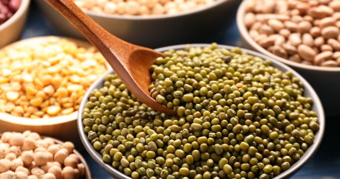 Taking peas with spoon from bowl, different types of legumes on blue wooden table, closeup