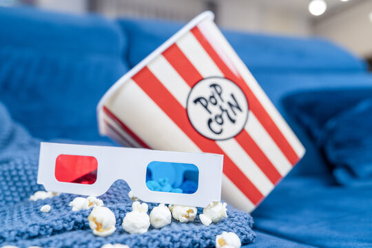 Red and blue cinema 3d glasses beside a popcorn bucket on a soft blue blanket, cozy movie night entertainment flat lay