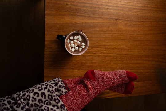 Top view of a cozy hot chocolate mug on a wooden table with feet in red socks
