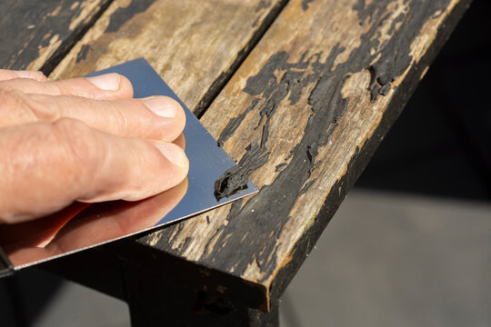 Man scraping loose peeling black paint off a wooden outdoor patio table. Garden DIY chore concept.